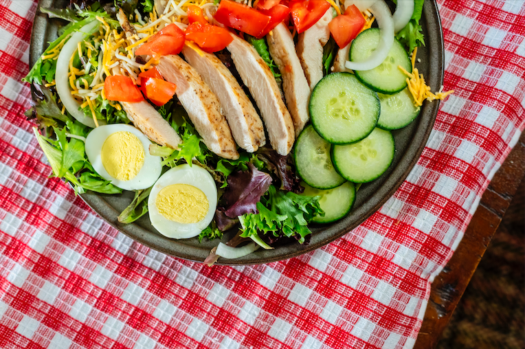 A salad with grilled chicken, sliced cucumbers, eggs, and tomatoes on a red gingham tablecloth.